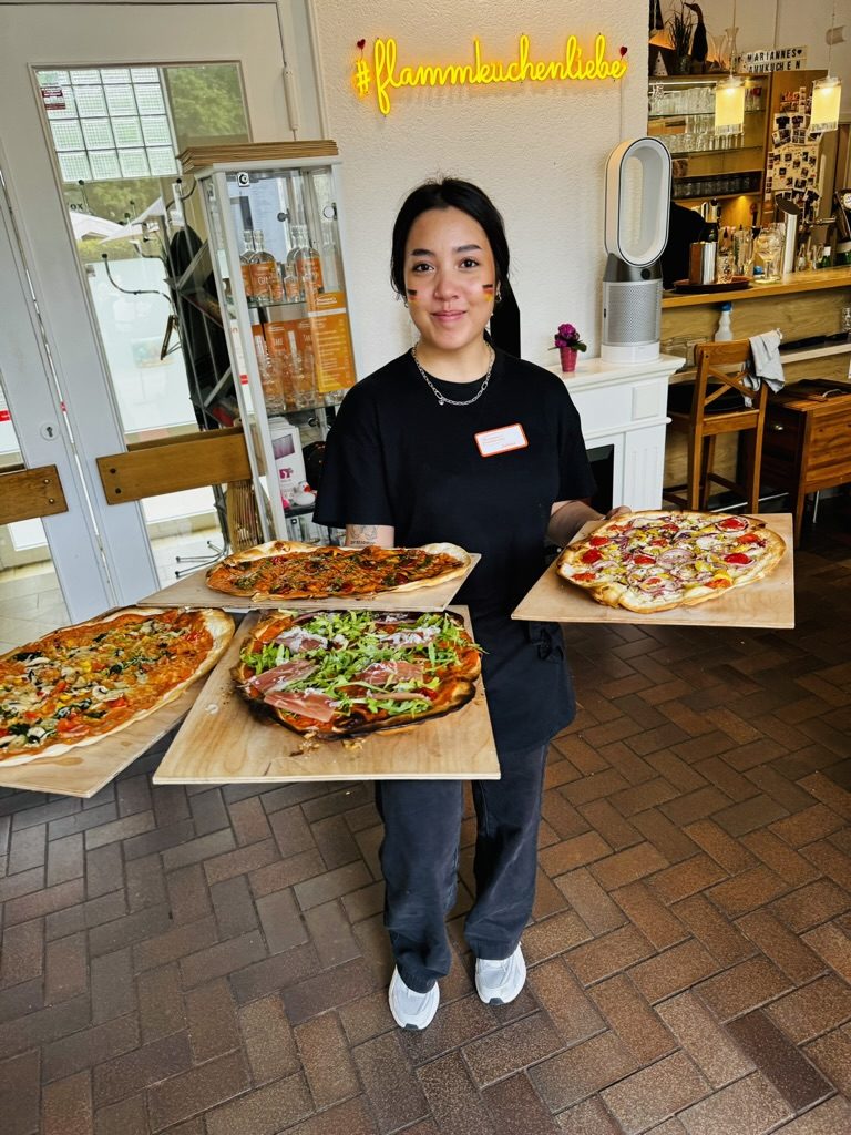 Eine Servicekraft balanciert vier Flammkuchenbretter auf beiden Armen, darüber die Neonschrift #flammkuchenliebe