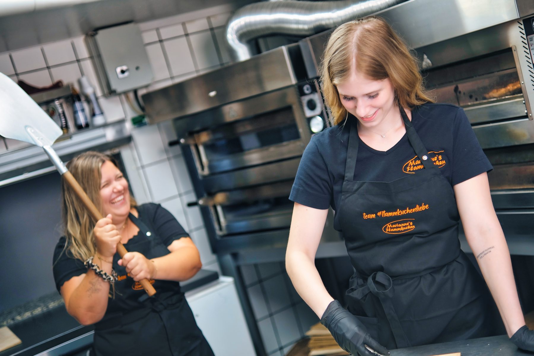 Zwei Mitarbeitende in der Küche vor den Öfen, eine mit dem Flammkuchenschieber in der Hand