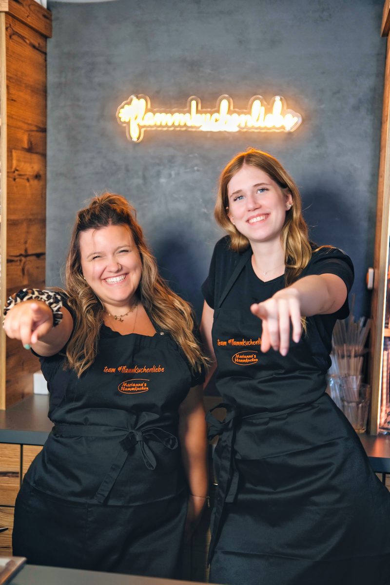 Zwei Mitarbeitende in Team-Schürzen zeigen lachend in die Kamera, darüber die Neonschrift #flammkuchenliebe