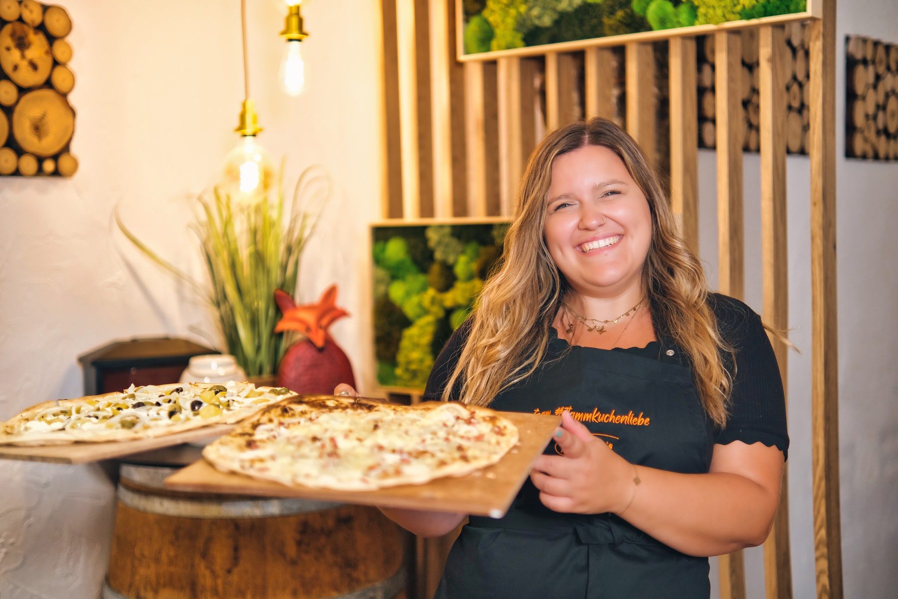 Ein Teammitglied trägt zwei fertige Flammkuchen auf Holzbrettern durch den Gastraum